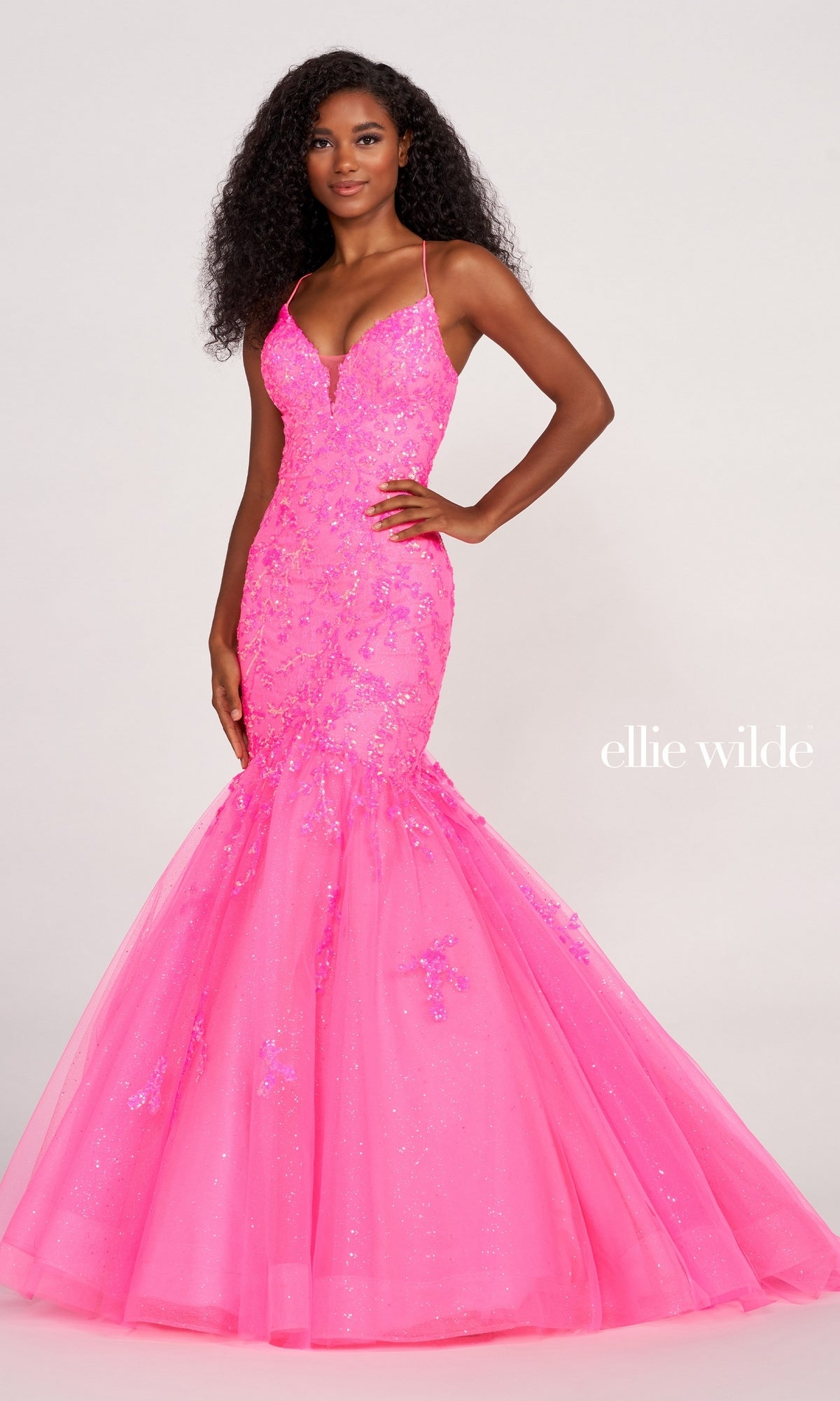 A woman poses in an Ellie Wilde Glitter-Tulle Long Mermaid Prom Gown featuring bright pink sequins, floral embellishments, thin straps, and a flared glitter tulle skirt against a plain light background.