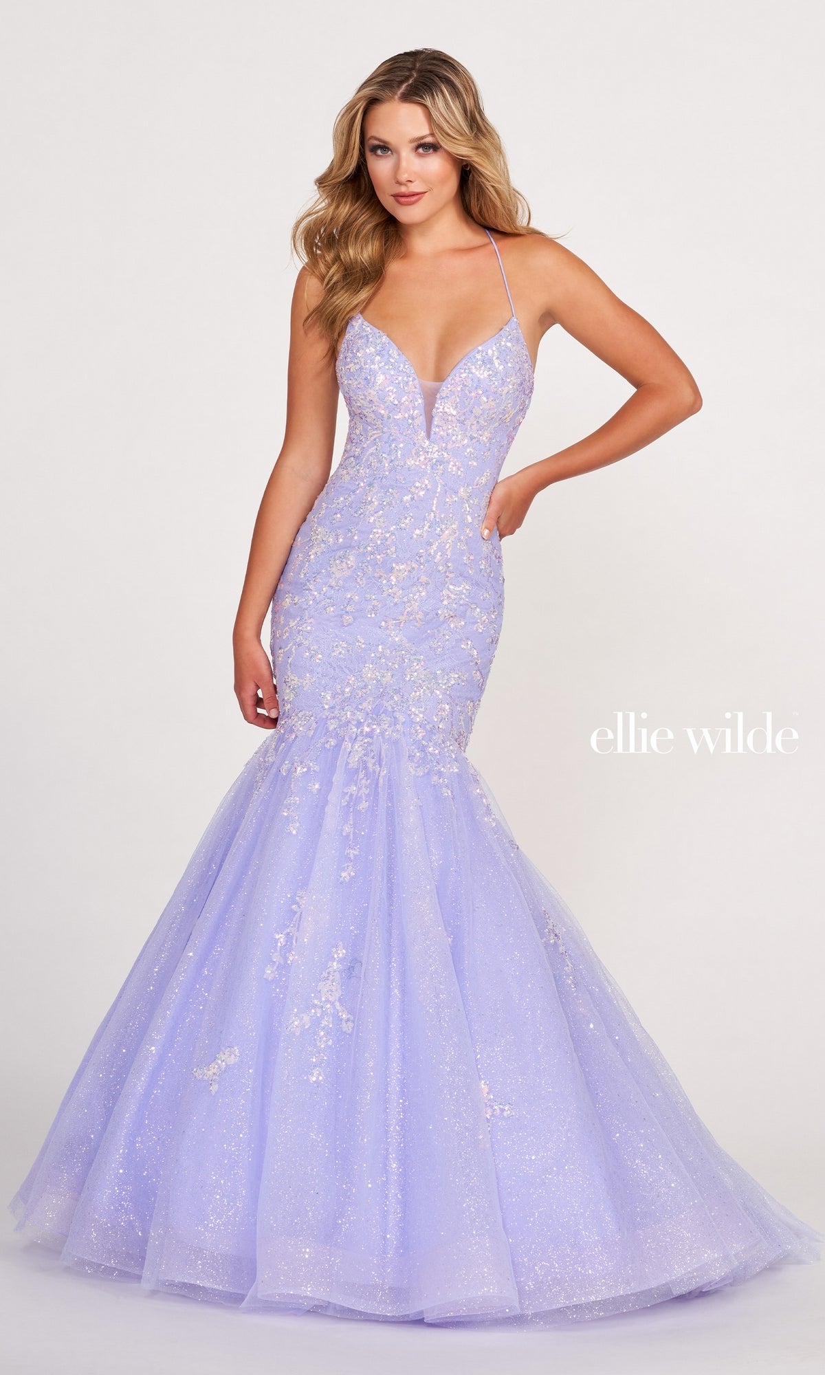 A woman stands against a plain background wearing the Ellie Wilde Glitter-Tulle Long Mermaid Prom Gown in light purple with thin straps. The text 