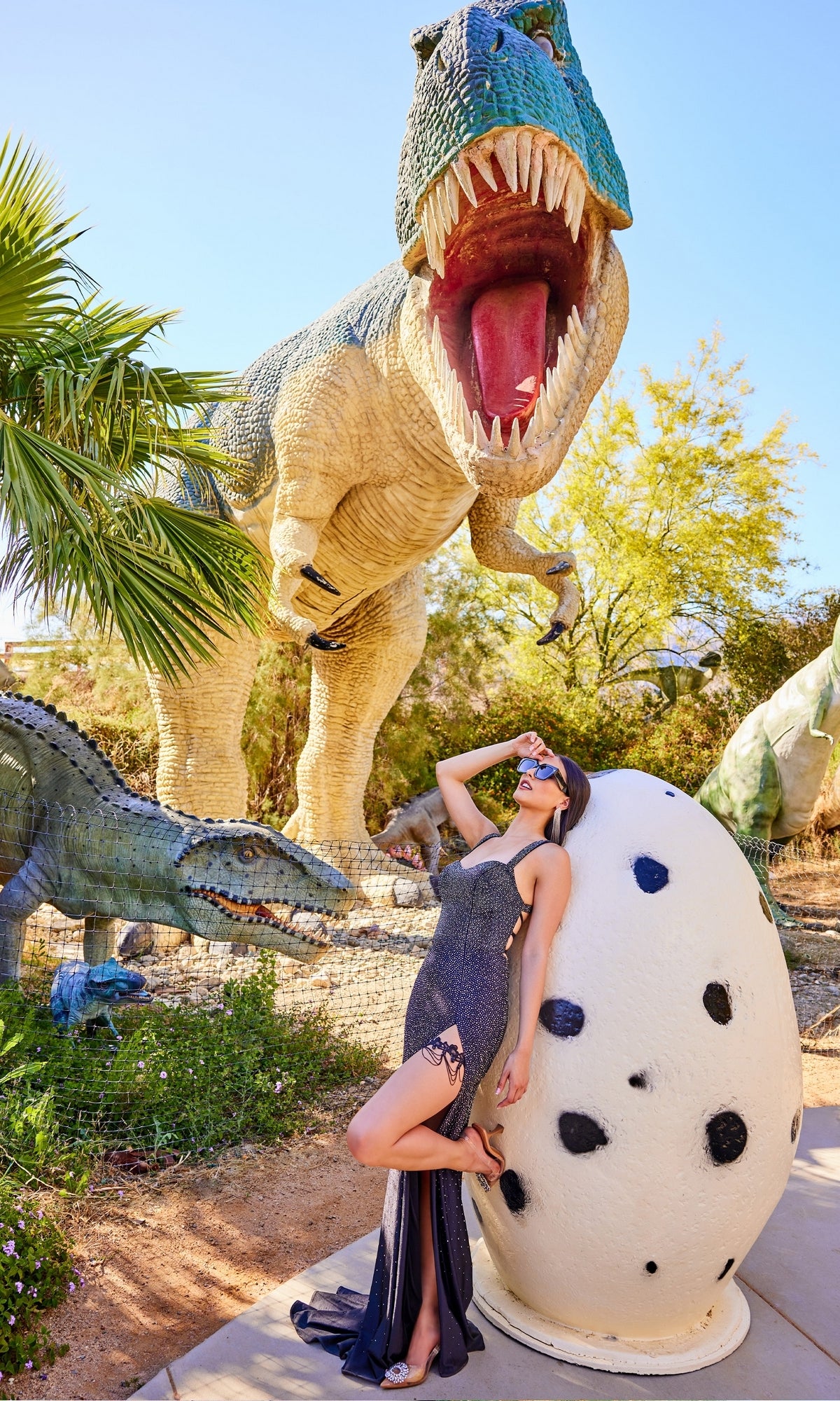 A woman wearing the Strappy-Back Long Beaded Prom Dress with Garter poses by a large, white, spotted dinosaur egg, with T-rex and other dinosaur statues in the sunny outdoor background.