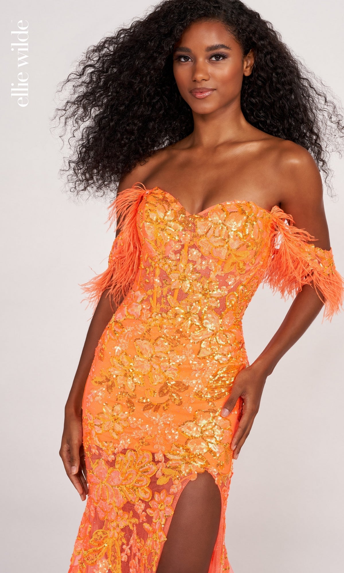 A woman poses confidently against a light background in the Feather-Trim Off-Shoulder Long Sequin Prom Dress—a bright orange gown with sequins, feathered sleeves, and a thigh-high slit.