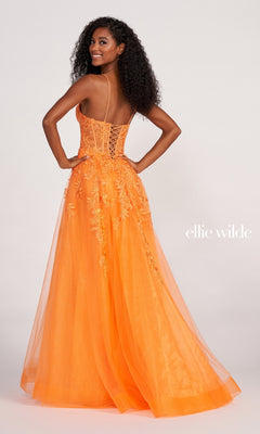 A woman in the Sheer-Corset Long Lace Prom Ball Gown EW34036, featuring orange lace with a fitted bodice and thin straps, stands facing away and smiles over her shoulder. The "Ellie Wilde" logo appears on the right.