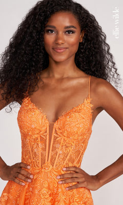 A woman with long curly hair poses with her hands on her hips against a light background, wearing the Sheer-Corset Long Lace Prom Ball Gown EW34036 by Ellie Wilde, featuring orange embroidery and spaghetti straps with a fitted bodice.