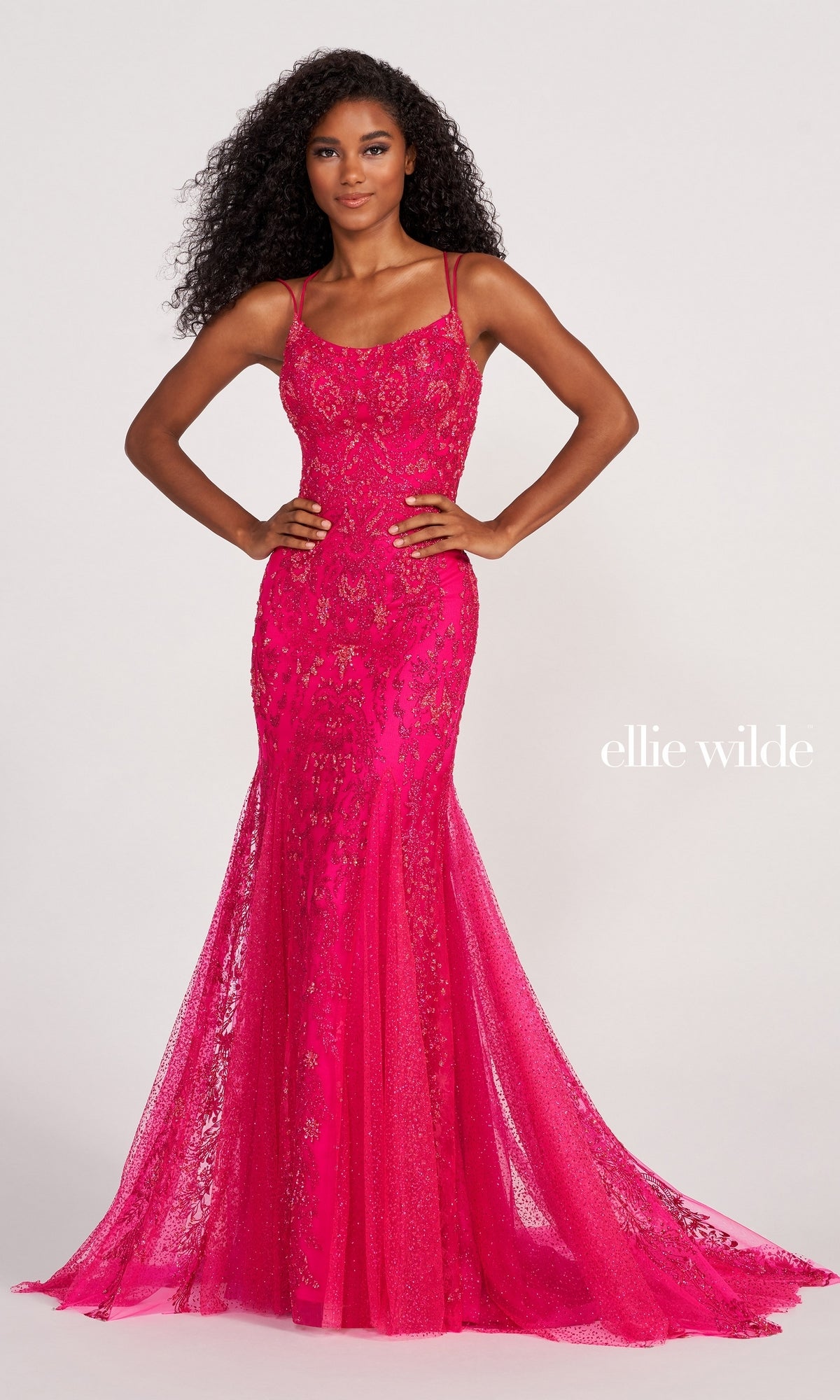 A woman wears the Glitter-Jersey Long Mermaid Ellie Wilde Prom Gown, a floor-length, sleeveless pink dress with sequin details. The Ellie Wilde brand logo is visible on the right side.