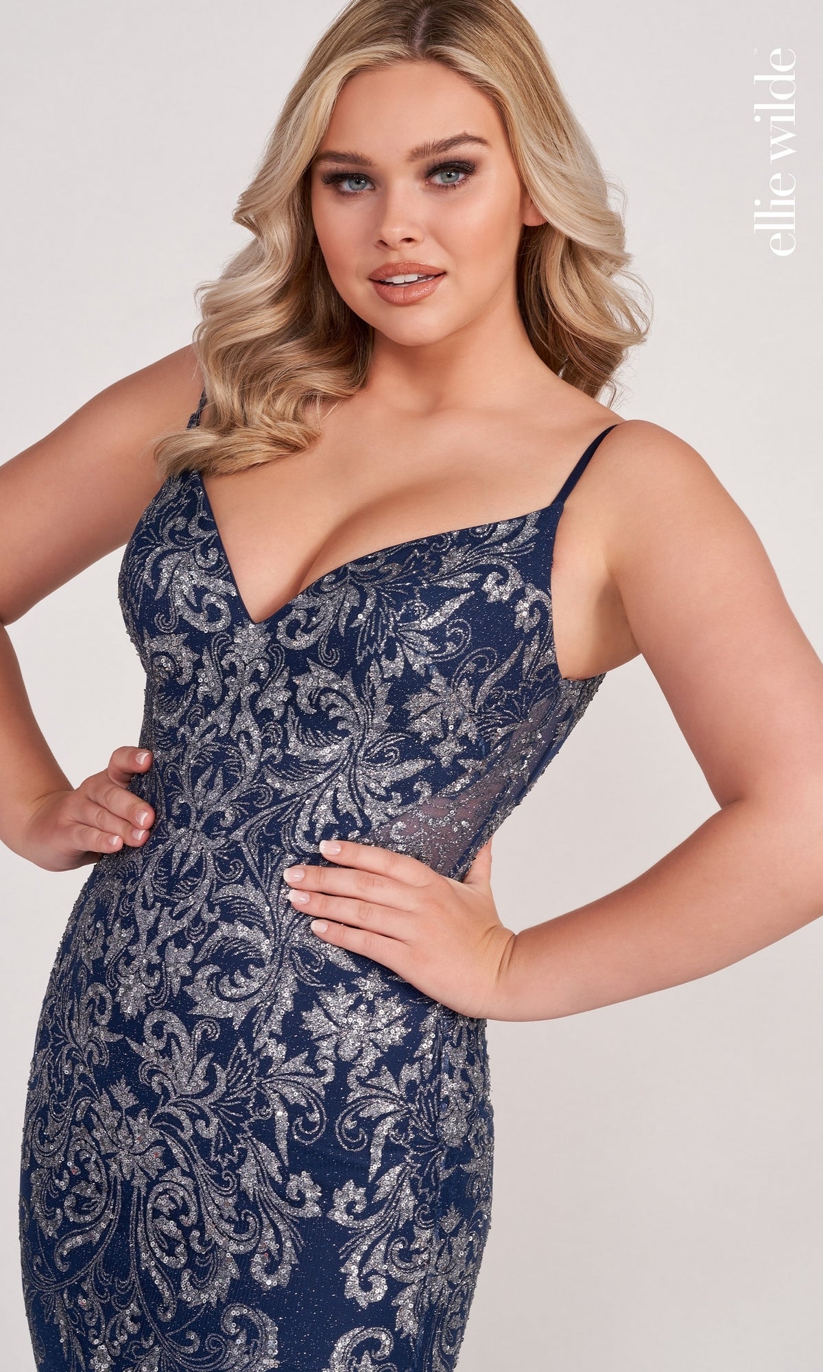A woman poses with one hand on her hip in the Sequin and Glitter Mermaid Prom Dress by Ellie Wilde, a navy blue gown adorned with silver patterns, against a light background. 