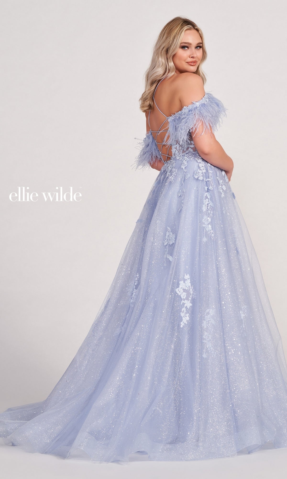 A woman poses with her back turned in the Glitter Ball Gown With Off The Shoulder Feather Sleeves, showcasing its elegant lace-up corset back and sparkling lace details.