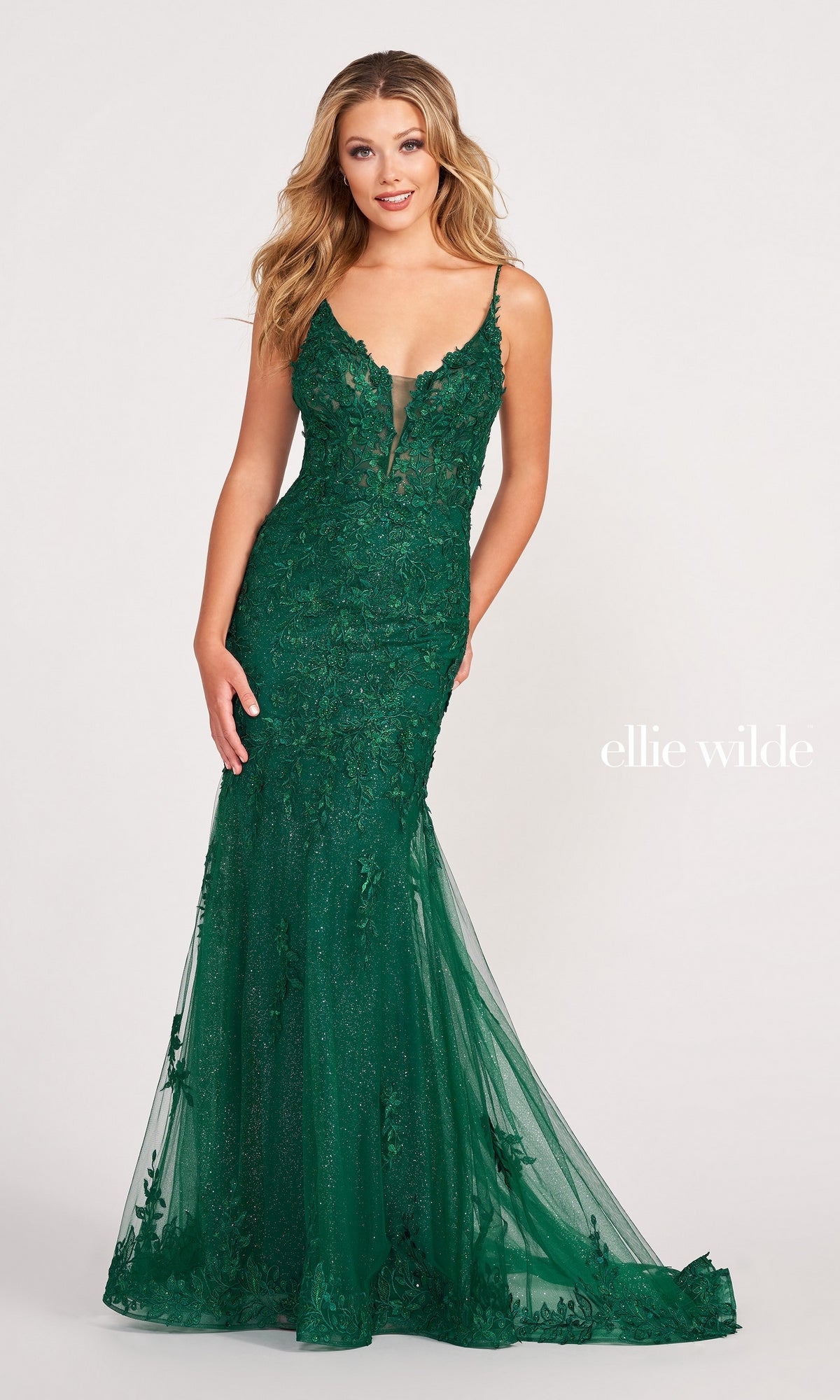 A woman wears the Lace Embroidered Mermaid Gown With Sheer Bodice by Ellie Wilde—a fitted, floor-length green lace dress with a deep V neckline and sheer corset details—standing against a plain light background with 