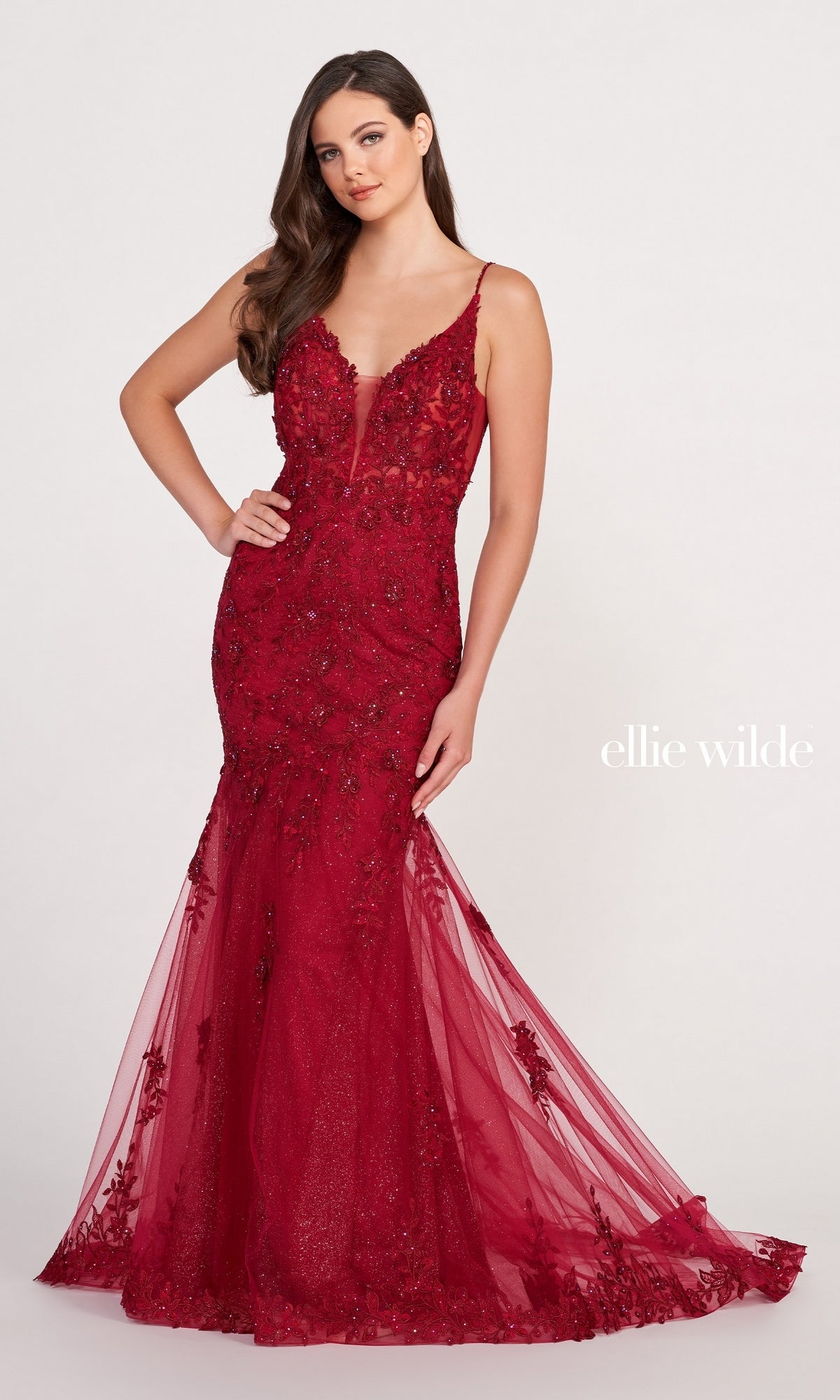 A woman stands against a plain background in the Lace Embroidered Mermaid Gown With Sheer Bodice, featuring deep red lace, floral embellishments, a sheer corset bodice, and a tulle train. The 