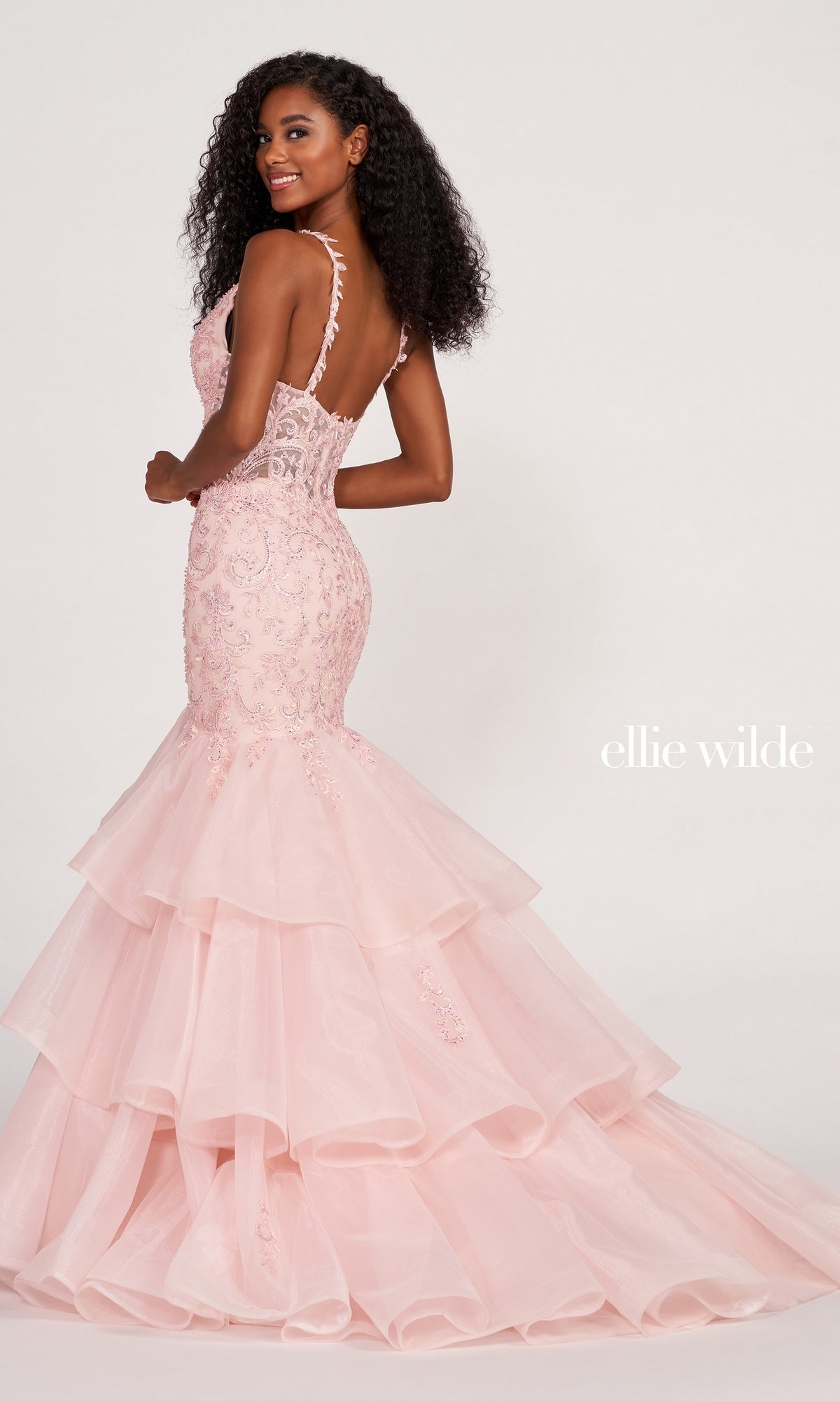 A woman models the Ellie Wilde EW34082 Layered Skirt Mermaid Gown in light pink, featuring thin straps, a low back, ruffled hem, and delicate embroidered details against a plain light background.