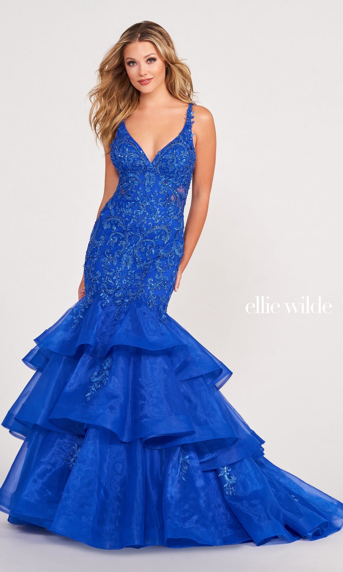 A woman wears the Ellie Wilde Gown EW34082, a royal blue, sleeveless mermaid dress with floral embroidery and a layered skirt, standing against a light background that highlights its elegant design.