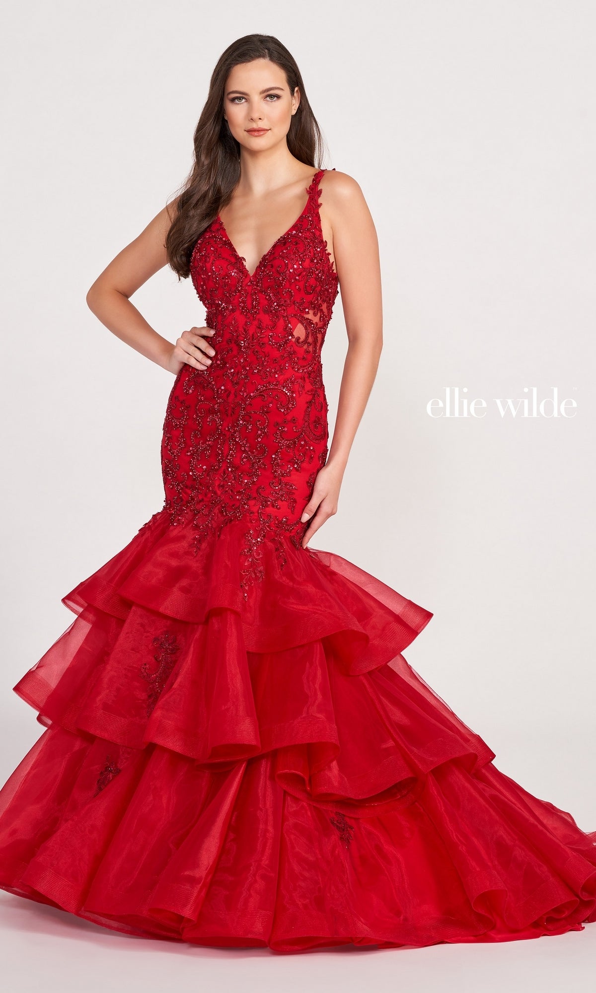 A woman poses in the Layered Skirt Mermaid Ellie Wilde Gown EW34082, a red dress with lace, bead detailing, and a dramatic ruffled hem, against a plain white background. 