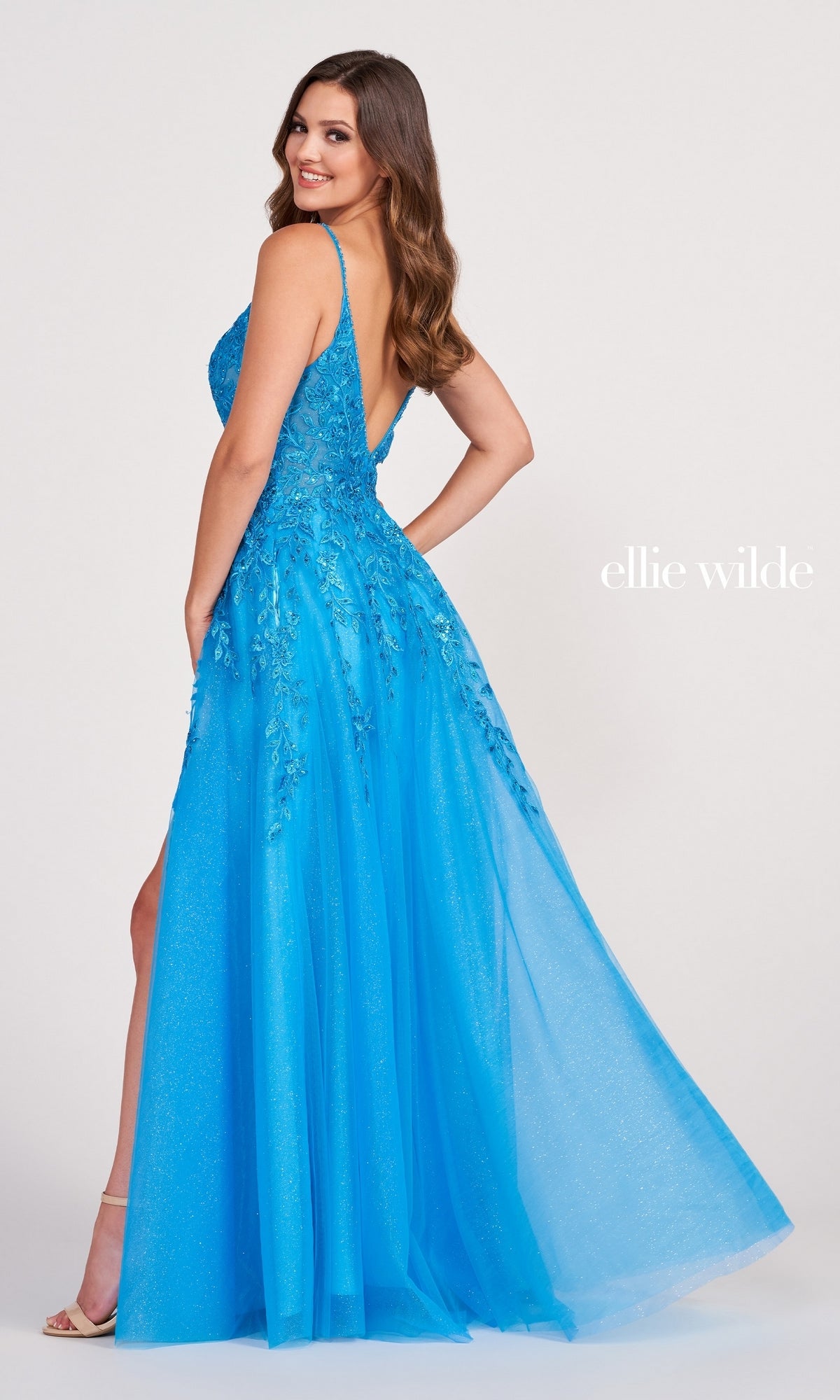 A woman models the Glitter-Tulle Long Ellie Wilde Prom Dress EW34089, a bright blue A-line gown with crystal beading, posing sideways and looking over her shoulder. The words 