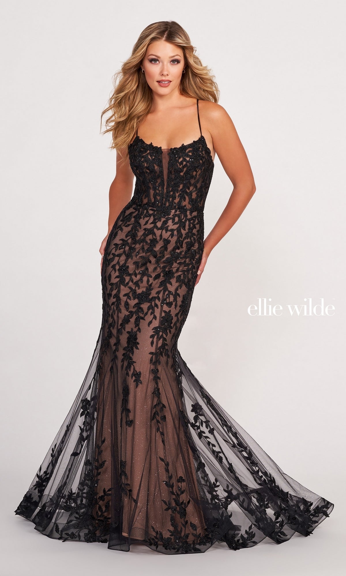 A woman wears the EW34090 Lace Applique Over Glitter Jersey Gown—floor-length, fitted, with black floral lace and thin straps—against a plain background. 