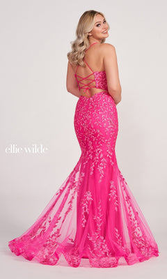 A woman wearing the Lace Applique Over Glitter Jersey Gown EW34090 in bright pink, featuring a mermaid silhouette, embroidered lace appliqué, and a lace-up open back, stands looking over her shoulder.