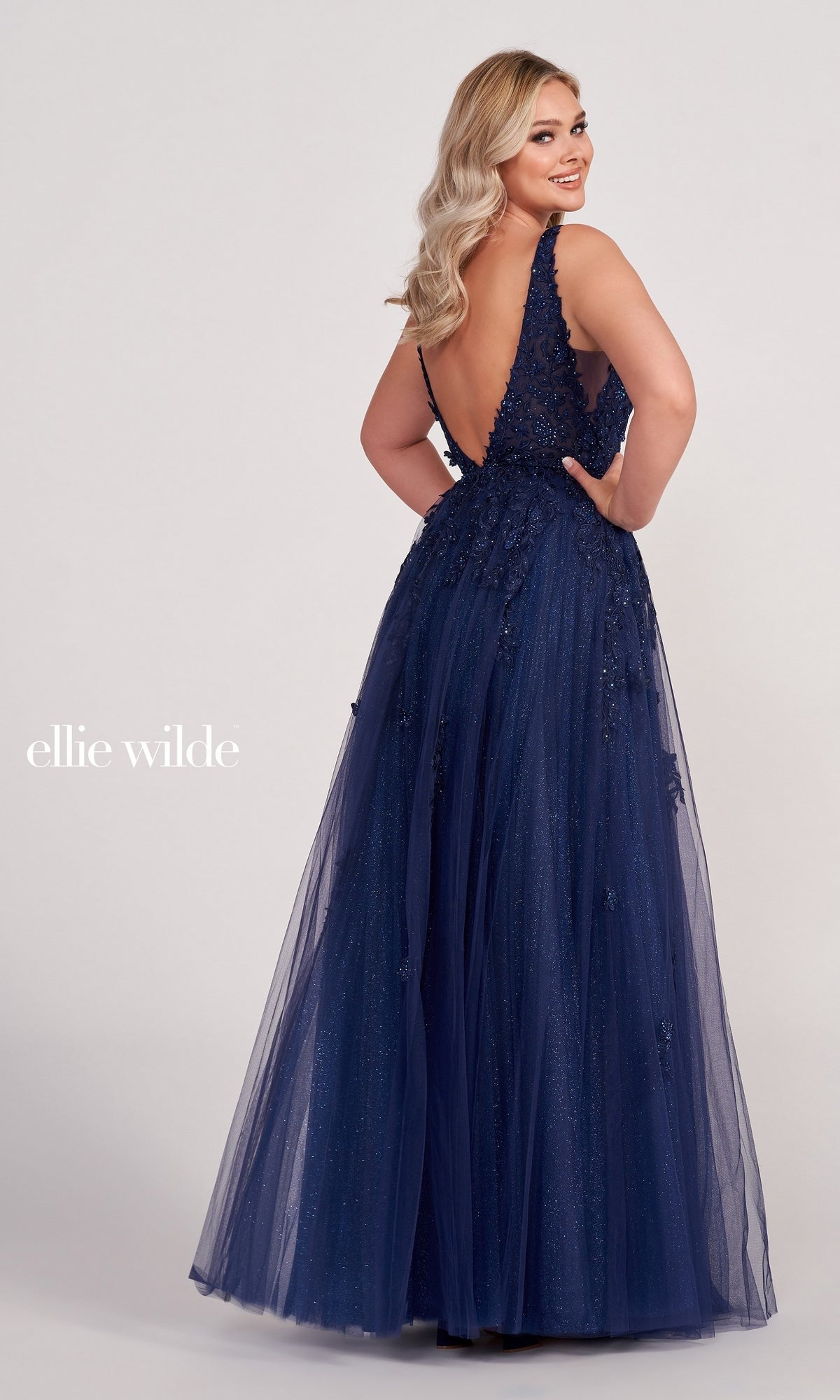 A woman poses in the Ellie Wilde Side-Slit Long Prom Ball Gown EW34103, a navy blue sleeveless evening dress with a deep V-back, floral embellishments, side slit, and a flowing tulle skirt.