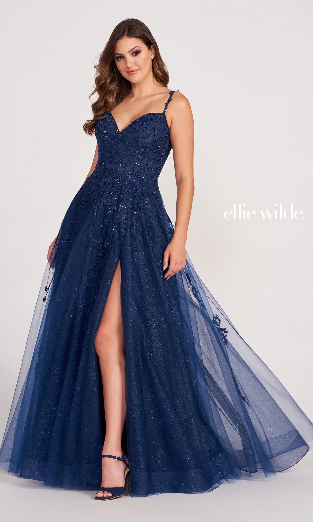 A woman wears the Ellie Wilde Long Glitter-Tulle A-Line Formal Dress in navy blue, featuring a thigh-high slit and sparkling details, standing before a plain light background. The text 