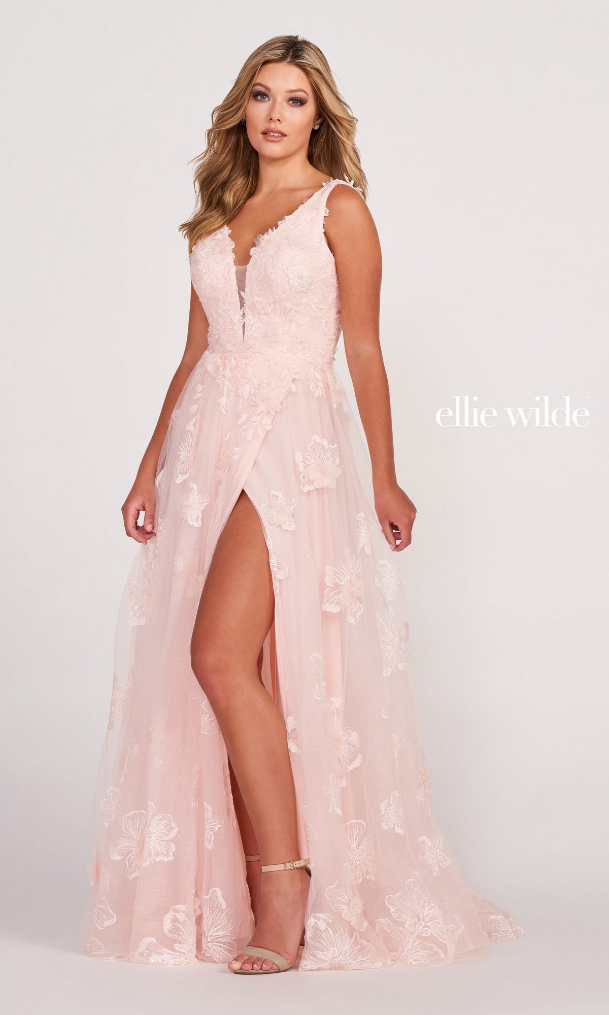 A woman wears the Ellie Wilde Faux-Wrap Embroidered-Lace Prom Dress—sleeveless, blush pink, with a deep V-neckline and high front slit—while standing against a plain background.