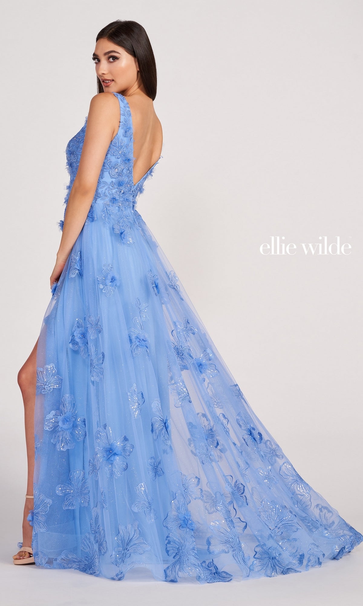 A woman poses in the Ellie Wilde Faux-Wrap Embroidered-Lace Prom Dress, featuring light blue lace, a deep V-back, high slit, and sheer overlay. The plain background displays the ellie wilde logo.