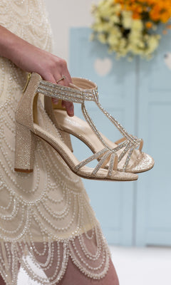 A person in a beaded dress holds Champagne Rhinestone Prom Shoes by Touch Ups 4527 featuring 2.75-inch glittery, strappy heels. Blue doors and yellow flowers appear in the background.