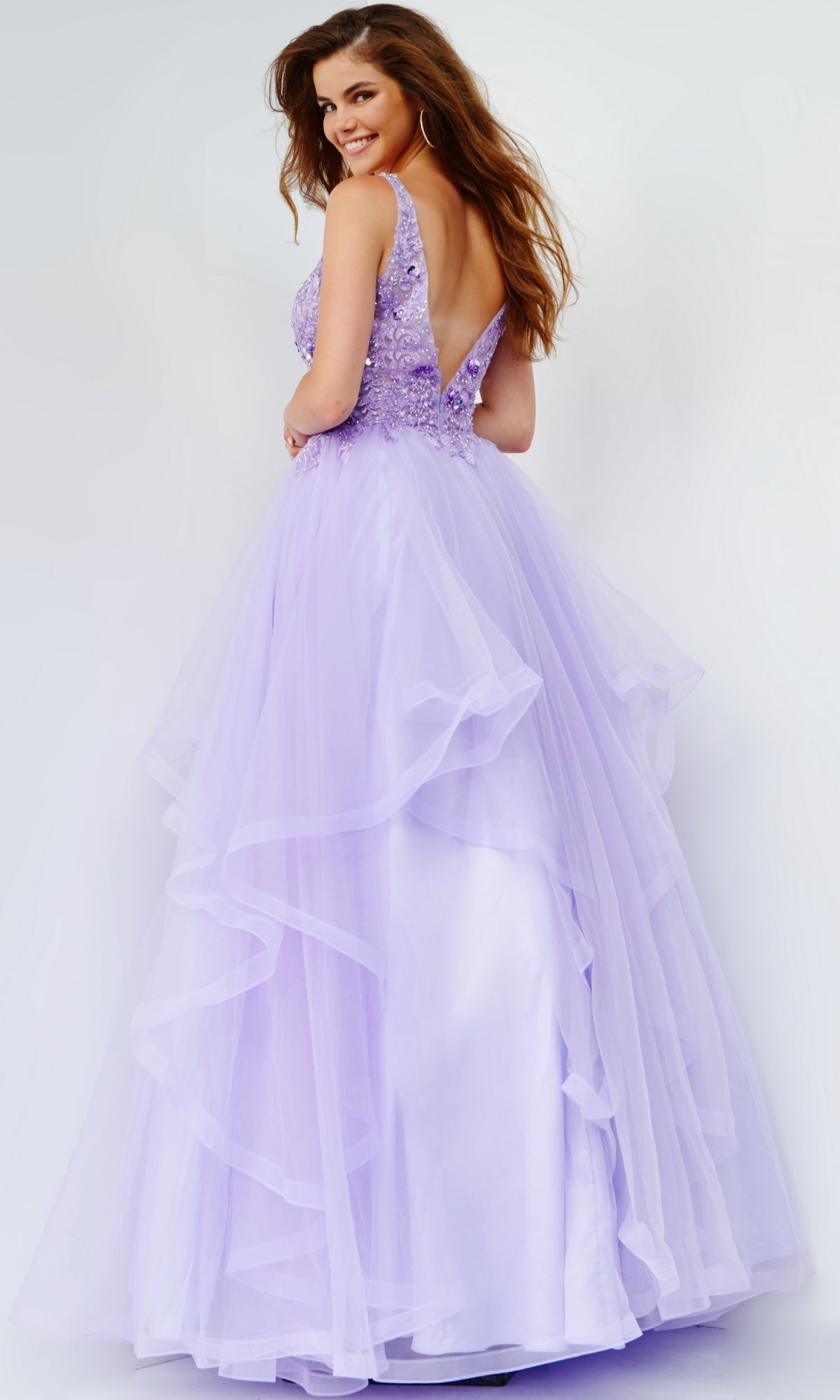 A woman smiles over her shoulder in a sleeveless lavender Jovani Prom Ball Gown with a layered tulle skirt and low back.