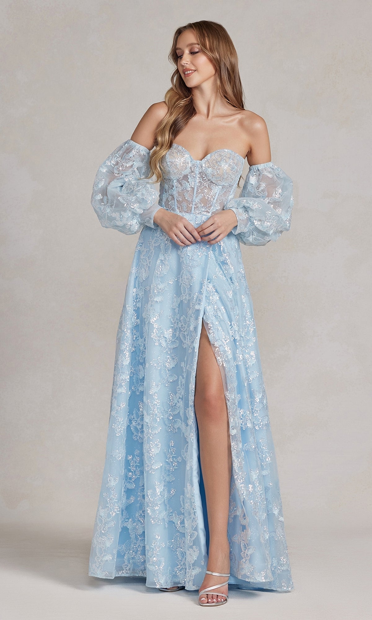 A woman wears the Long Puff-Sleeve Prom Dress with Sheer Bodice: a light blue, floral patterned A-line gown featuring sheer removable puff sleeves, a corset-style bodice, and a thigh-high slit.