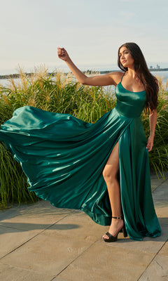 A woman stands outdoors near tall grass, holding out part of her Long Open-Back Formal Prom Dress by Faviana—an elegant emerald green gown with a thigh-high slit.