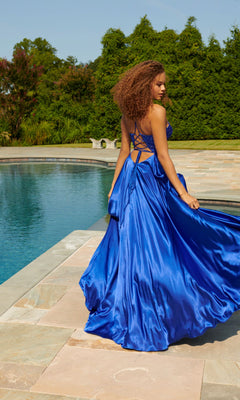 A woman in the Faviana Beaded-Bodice Long Prom Dress S10870 stands by a pool, holding her bright blue a-line gown with a beaded v-neck bodice, framed by lush greenery.