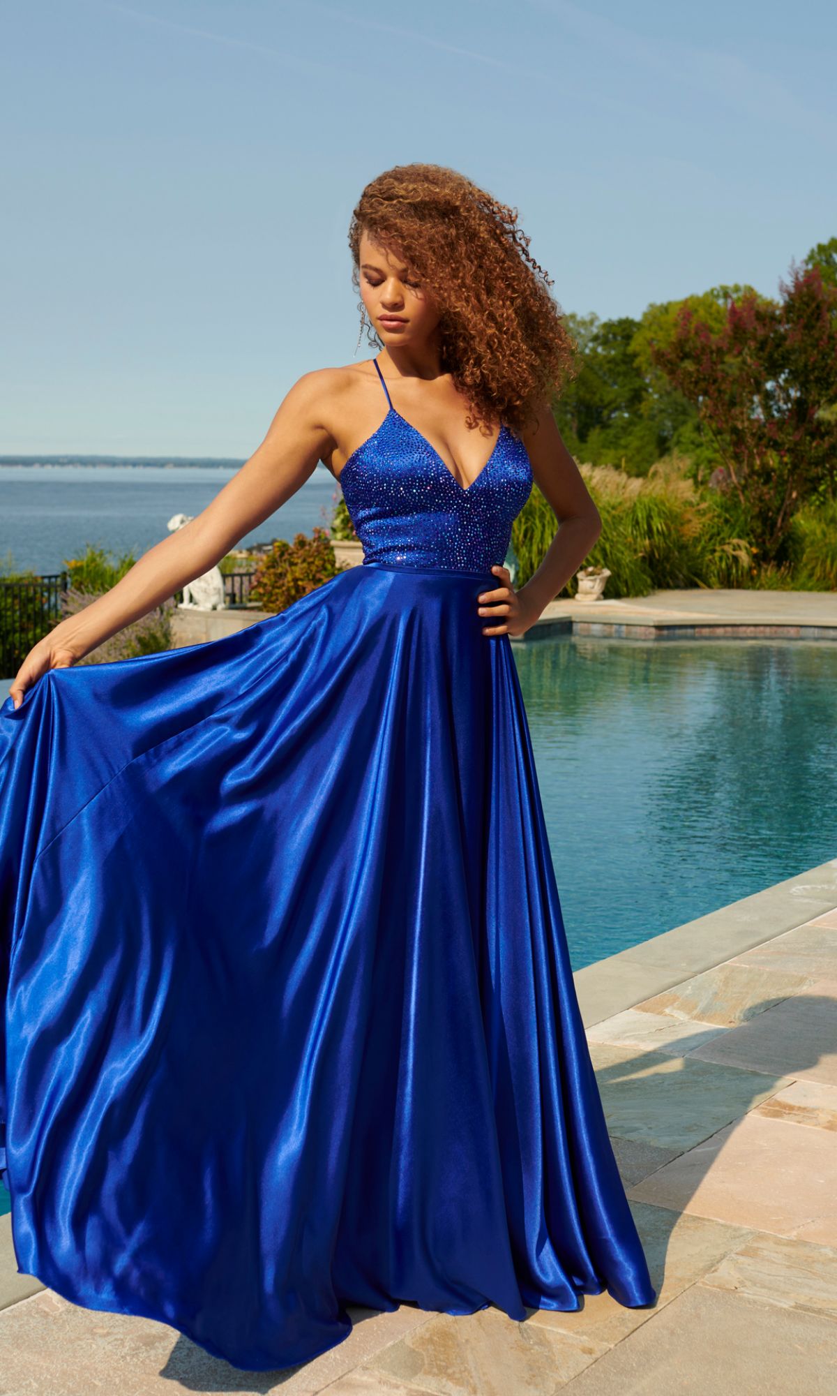 A woman stands by a pool in the Faviana Beaded-Bodice Long Prom Dress S10870, featuring a vibrant blue satin A-line skirt and beaded V-neck bodice. She holds out her elegant gown with trees and water in the background.