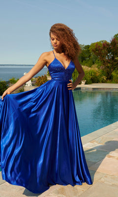 A woman stands by a pool in the Faviana Beaded-Bodice Long Prom Dress S10870, featuring a vibrant blue satin A-line skirt and beaded V-neck bodice. She holds out her elegant gown with trees and water in the background.