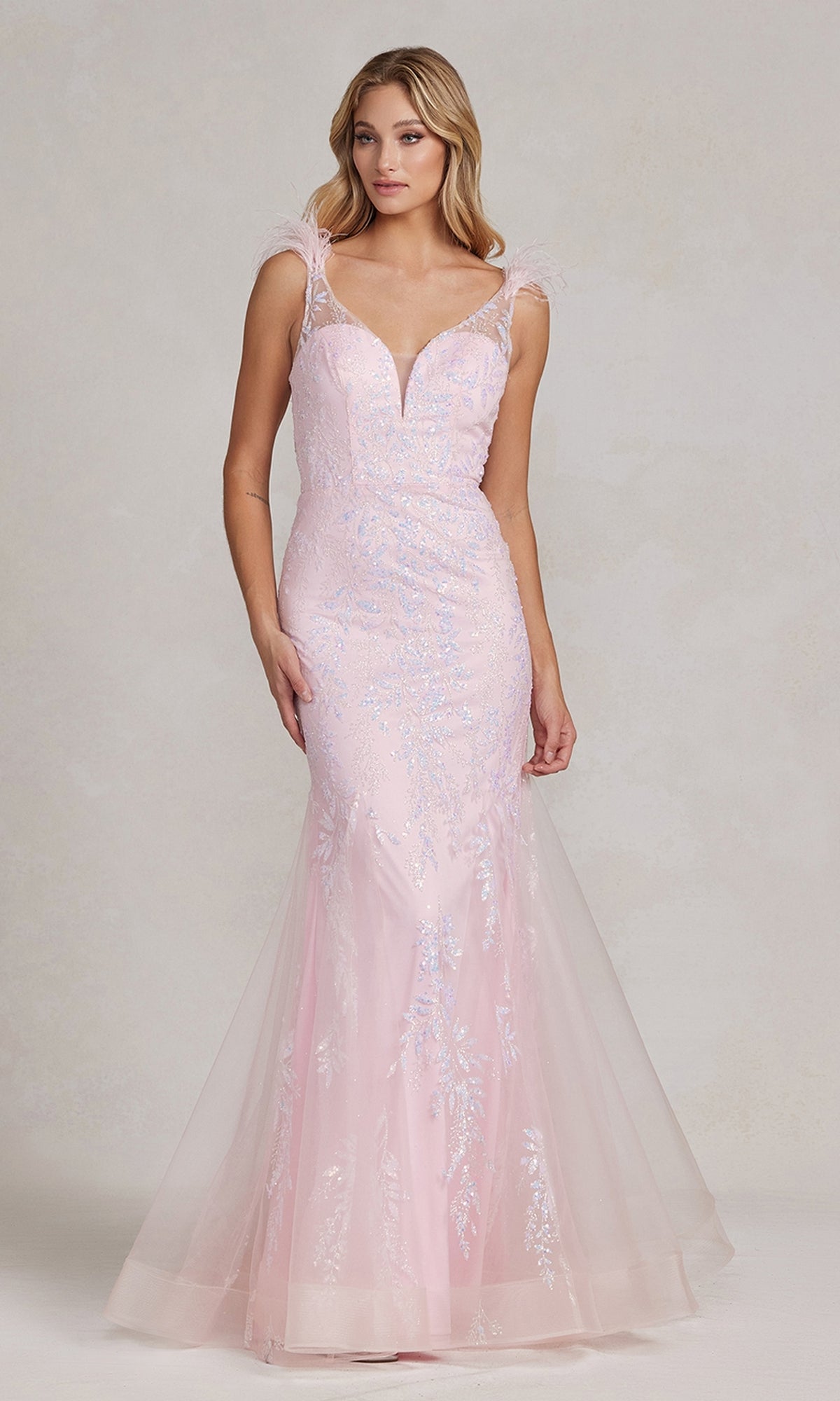 A woman stands against a neutral background wearing the Nox Anabel T1208 Feather-Strap Long Prom Dress in pink, featuring sheer feather straps and delicate floral embellishments.