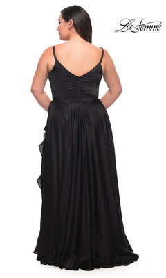 A woman models the Long Plus-Size La Femme Prom Dress, a floor-length black satin gown with a V-back and ruched details, featuring a ruffle hem. She stands facing away from the camera, with the La Femme logo in the upper right corner.