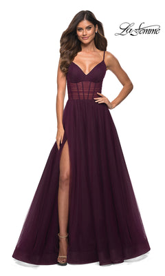 Woman modeling a Dark Berry Long La Femme Prom Dress with a sheer corset waist, thin straps, and high slit, standing before a white background featuring the "La Femme" logo.